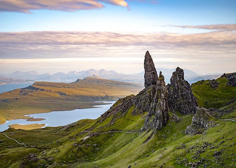 The Old Man of Storr rock pinnacles on the Isle of Skye at dawn with lochs, green ridges, and misty Cuillin mountains in the distance