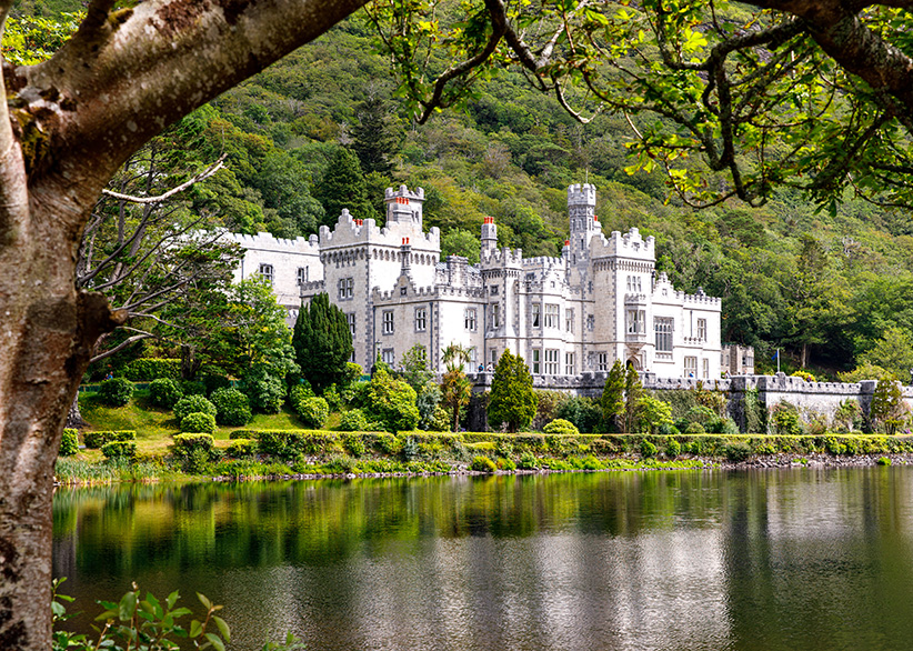 Kylemore Abbey framed by overhanging tree branches, with the castle-like stone building reflected in the lake and lush green woodland rising behind it in Connemara, Ireland