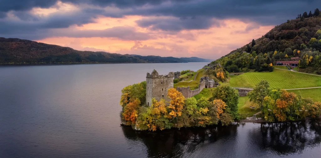 Panoramic aerial view of Urquhart Castle ruins on the shores of Loch Ness surrounded by autumn foliage with the loch stretching into the distance under a dramatic pink and purple sunset sky