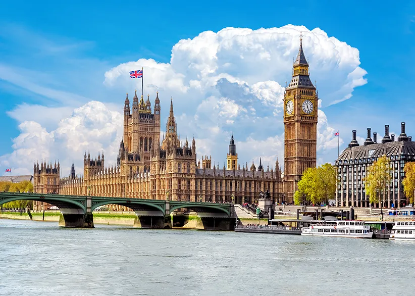 The Houses of Parliament and Big Ben seen from across the River Thames with Westminster Bridge, a Union Jack flag flying, and white clouds against a blue sky