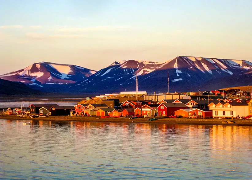 Longyearbyen's red and orange waterfront buildings glowing in the midnight sun with snow-streaked Arctic mountains reflected in the calm fjord, Svalbard, Norway