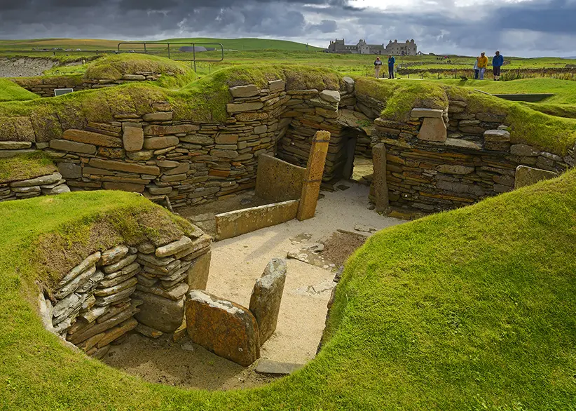 The ancient stone dwellings of Skara Brae Neolithic settlement with exposed rooms, stone furniture, and grass-covered walls, with visitors and green Orkney farmland in the background