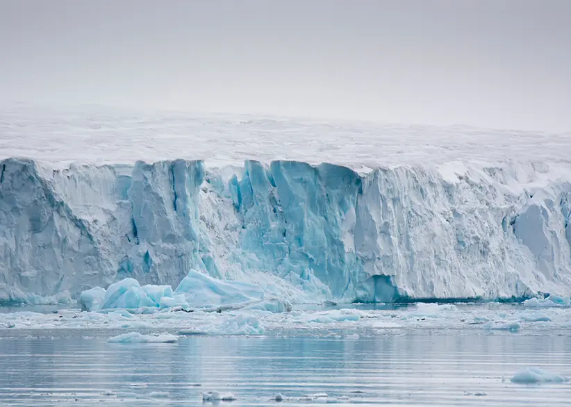 The towering blue ice wall of a glacier on Nordaustlandet with broken ice floating in the calm Arctic waters below under an overcast sky, Svalbard, Norway