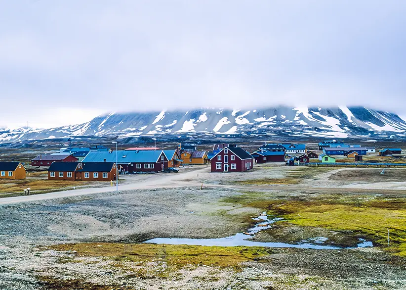 The remote Arctic research settlement of Ny-Ålesund with colourful wooden buildings on flat tundra and snow-capped mountains disappearing into low cloud, Svalbard, Norway