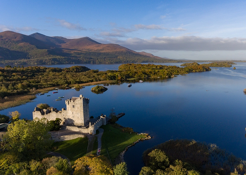 Aerial view of Ross Castle on the shores of Lough Leane in Killarney National Park with wooded islands, calm blue water, and the MacGillycuddy's Reeks mountains in the background