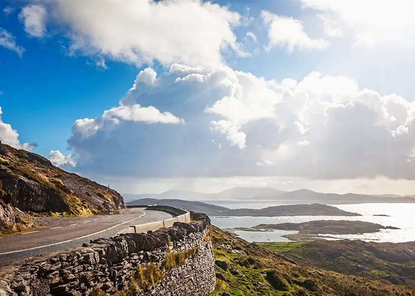 A winding coastal road on the Ring of Kerry with a stone wall, dramatic views over bays, islands, and mountains with sunlight breaking through the clouds, County Kerry, Ireland