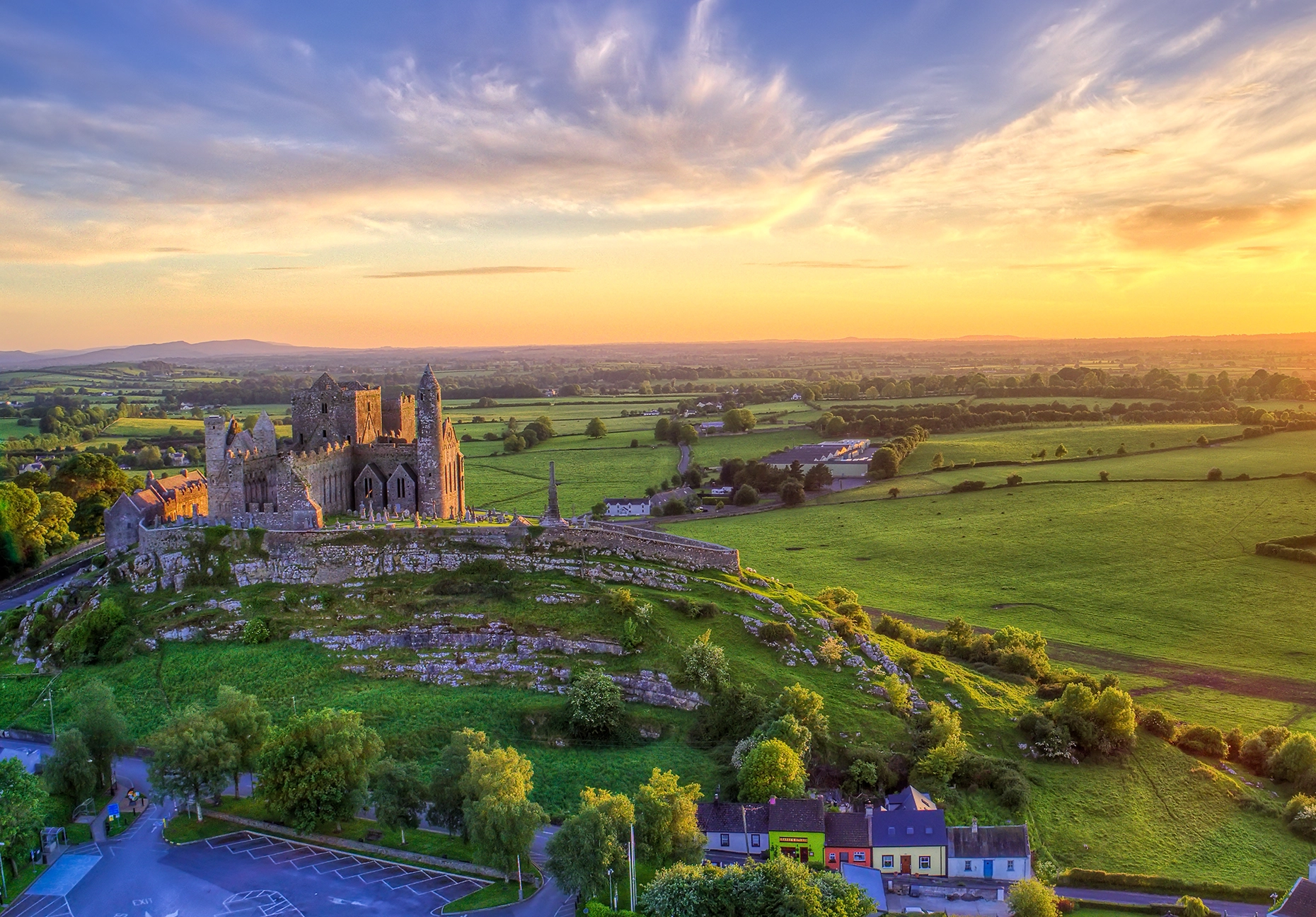 Aerial view of the Rock of Cashel at sunset with the medieval ruins glowing in warm light, the town of Cashel below, and rolling green Tipperary farmland stretching to the horizon