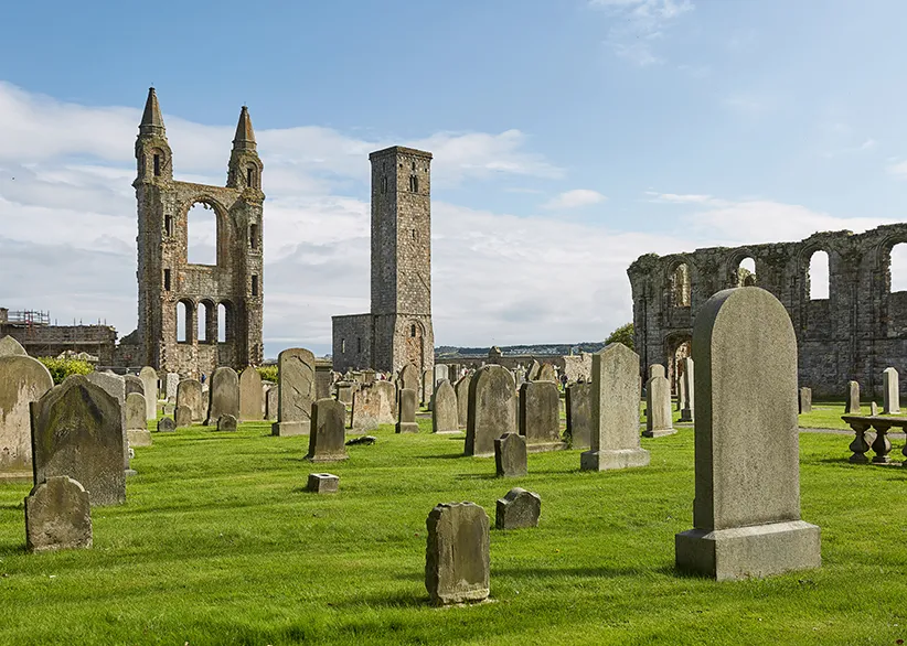 The ruins of St Andrews Cathedral with its remaining stone towers and arched walls surrounded by ancient gravestones on green lawns under a blue sky, St Andrews, Scotland