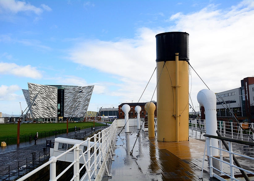 View from the deck of the SS Nomadic with its yellow funnel, looking towards the angular Titanic Belfast museum building in the Titanic Quarter on a rainy day