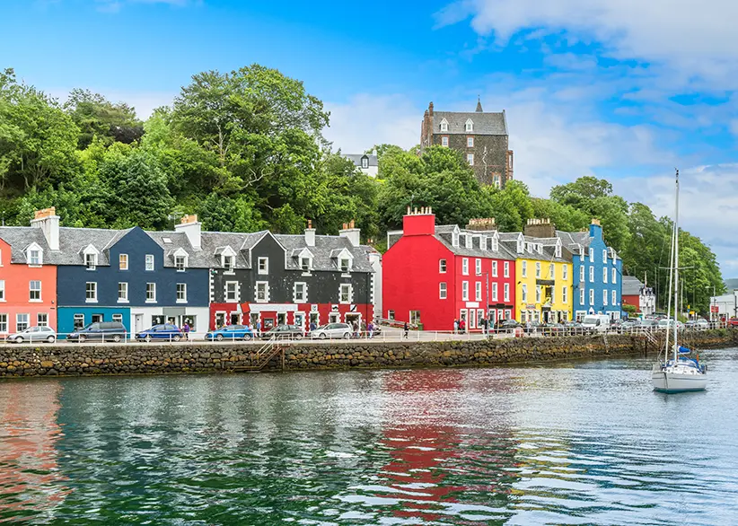 The colourful painted waterfront buildings of Tobermory harbour in red, blue, yellow, and grey with a sailing boat and green wooded hillside behind, Isle of Mull, Scotland