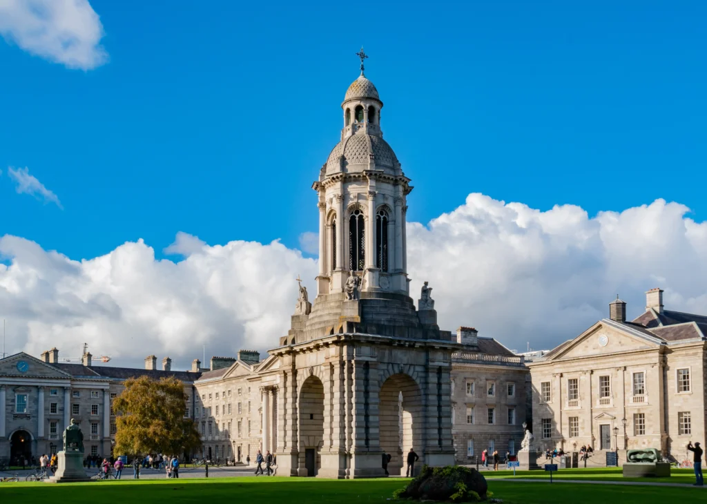 The Campanile bell tower in the cobbled courtyard of Trinity College Dublin on a sunny day with historic stone buildings, green lawns, and visitors walking around