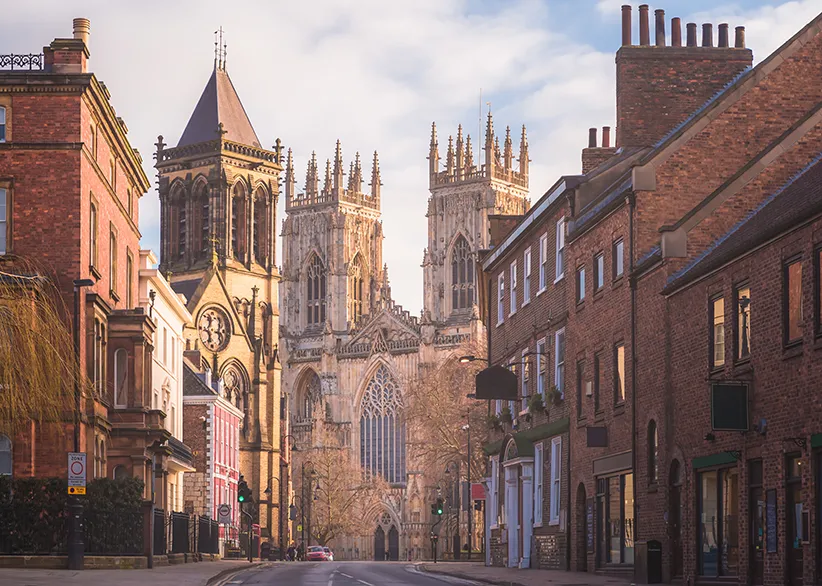 York Minster's Gothic twin towers rising above a quiet street lined with red-brick buildings in warm evening light, York, England