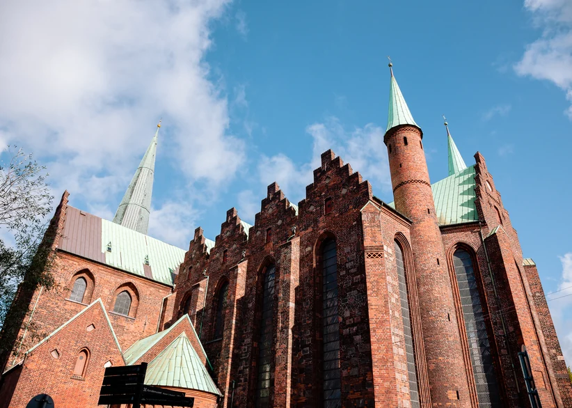 Low-angle view of Aarhus Cathedral's red-brick exterior, showing stepped gables, tall arched windows, a copper-green tower with a slender spire, and a taller spire visible in the background against a blue sky with white clouds.