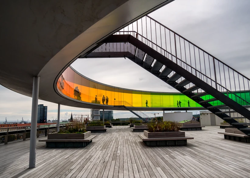 A spiral staircase on a rooftop terrace leads up to a circular walkway of rainbow-colored glass panels, with wooden decking, planters, and silhouettes of visitors walking through the colored glass in the background.