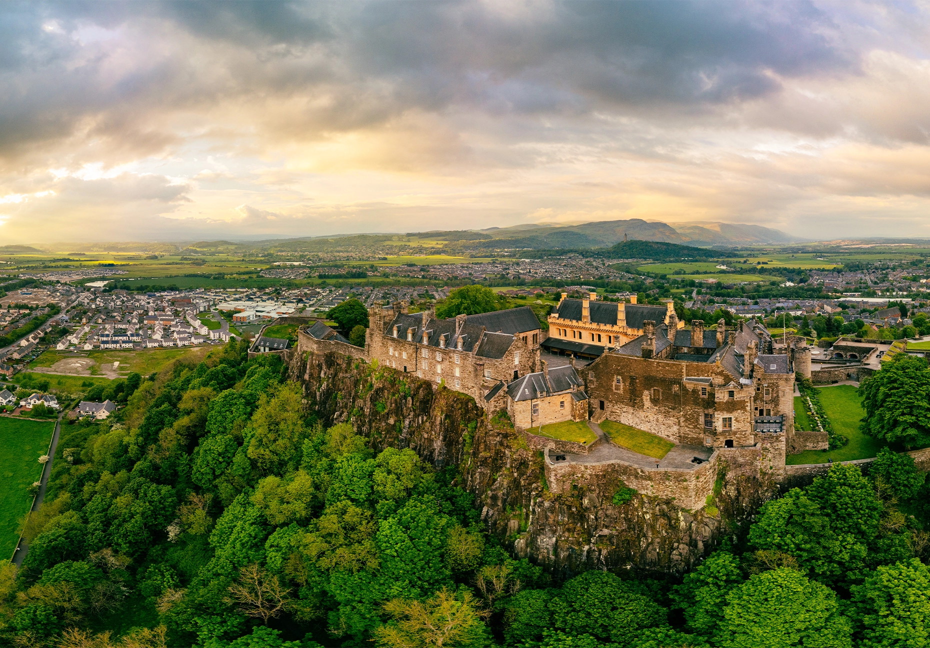 Stirling Castle sits atop a wooded crag above the town, with hills beyond at sunset.