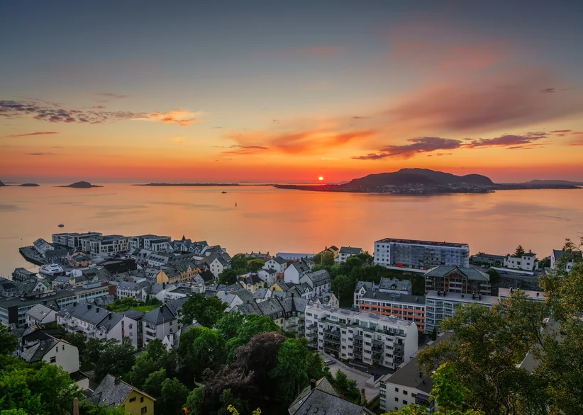 Aerial view of the coastal town of Ålesund at sunset, with rooftops and waterfront buildings in the foreground and a vivid orange and pink sky reflecting on the calm fjord waters, with distant islands silhouetted on the horizon.