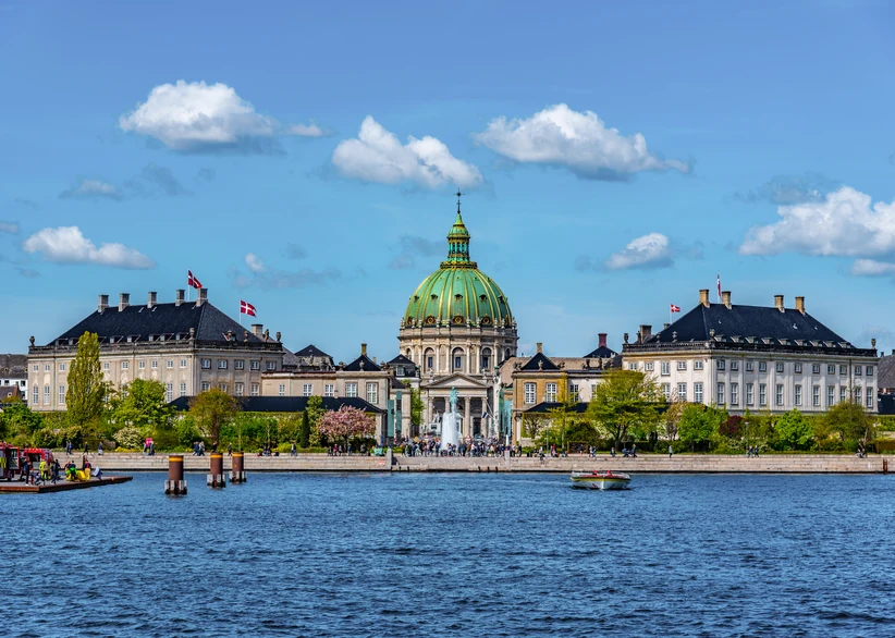 View across a blue harbor toward Frederik's Church's green copper dome rising above waterfront palace buildings, with Danish flags flying, small boats on the water, and a fountain and crowds along the promenade.