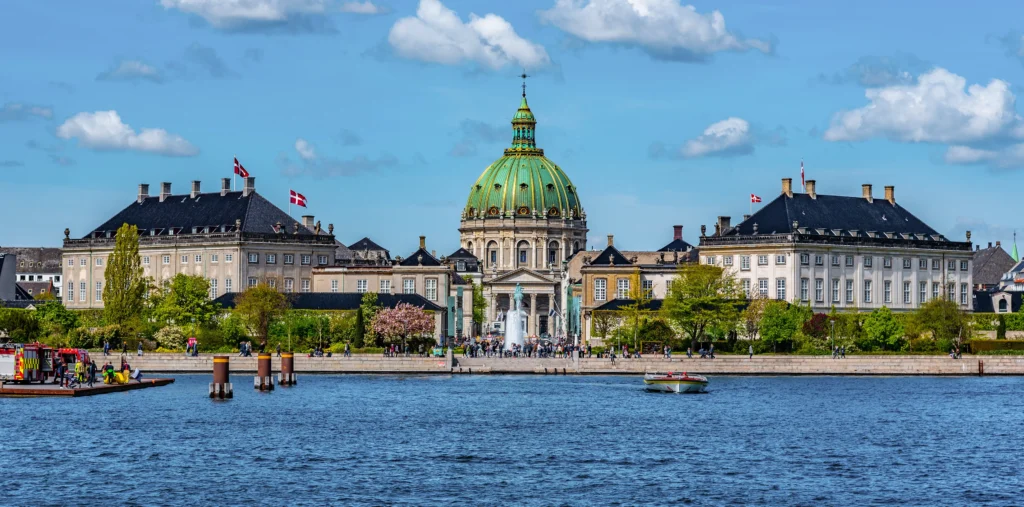 Frederik's Church's green copper dome rises behind the twin wings of Amalienborg Palace, seen across the blue harbor with a fountain and crowds gathered along the waterfront promenade, small boats on the water, and Danish flags flying
