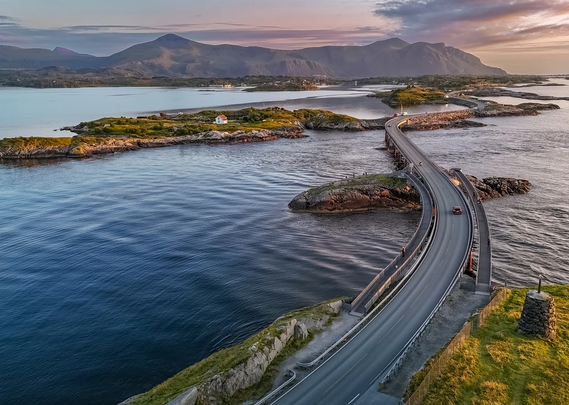 Aerial view of the Atlantic Ocean Road's curving bridge winding across small rocky skerries and islets, with a car crossing the causeway at sunset beneath layered mountains on the horizon.