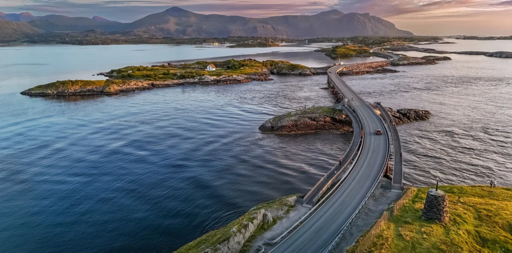Aerial view of the Atlantic Ocean Road's curving bridge winding across small rocky skerries and islets, with a car crossing the causeway at sunset beneath layered mountains on the horizon.