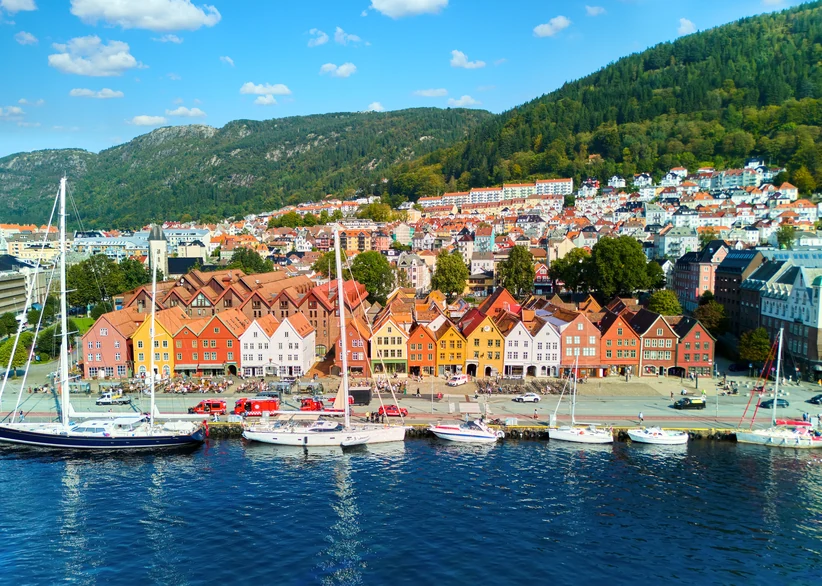 Daytime view of sailboats moored along Bergen's harbor with the historic wooden gabled buildings of Bryggen wharf lining the waterfront, backed by the city's colorful hillside houses and forested mountains under a partly cloudy sky.