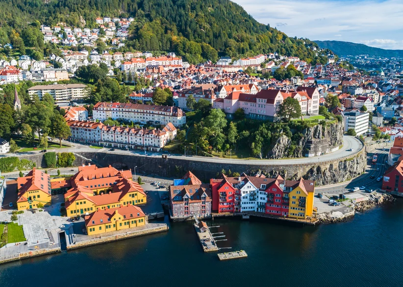 Aerial view of Bergen's waterfront, with red and yellow warehouse buildings along the harbor's edge, a rocky promontory topped with historic buildings, and the city's red-roofed houses spreading up a forested hillside.