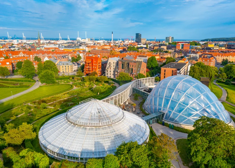 Aerial view of the domed glass greenhouses of the Aarhus Botanical Garden surrounded by green lawns and trees, with the red-roofed cityscape of Aarhus and its harbor cranes stretching to the coastline in the background.