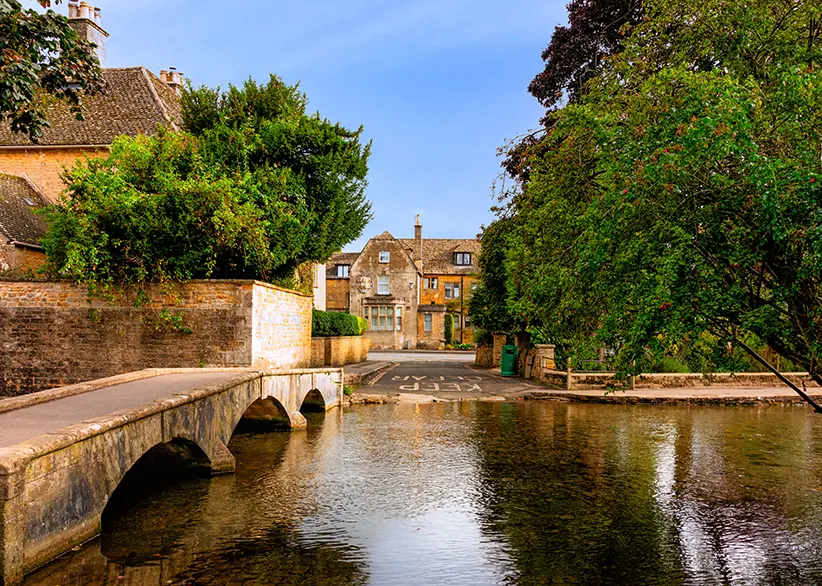 A low stone bridge over the River Windrush flowing through the honey-coloured Cotswold village of Bourton-on-the-Water with trees and stone cottages on a sunny day
