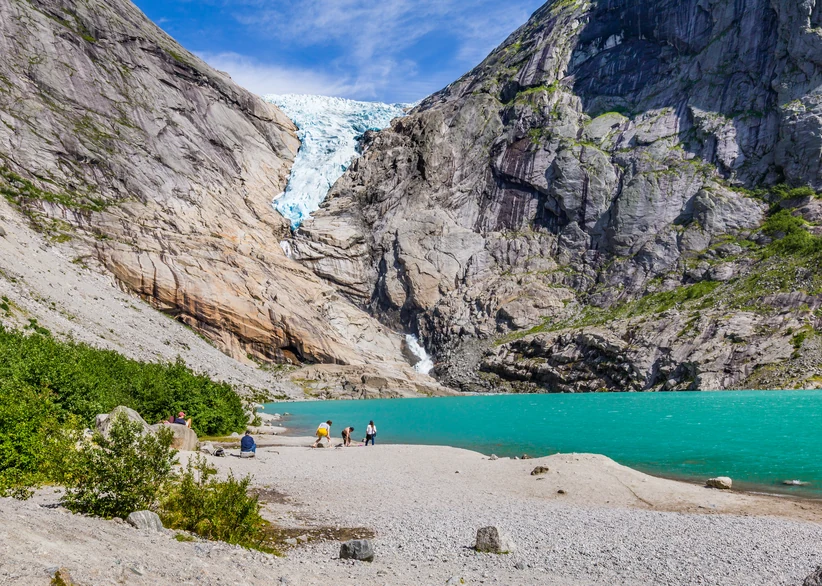 The Briksdalsbreen glacier tongue spills down a narrow rock gorge into a bright turquoise glacial lake, with visitors walking along a gravel beach in the foreground surrounded by steep, striated mountain walls.