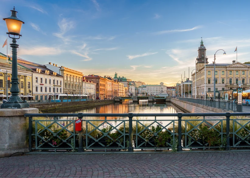 A tree-lined canal in central Gothenburg reflects historic facades and a tram at sunset, with an ornate green streetlamp and floral-decorated railing in the foreground and a clock tower building visible on the right.