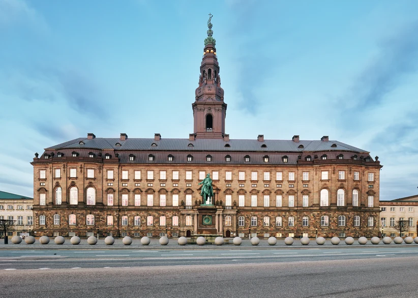 Twilight view of Christiansborg Palace's long sandstone facade with its tall central spire, an equestrian statue in front of the entrance, and a row of round stone bollards lining the empty street at dusk.