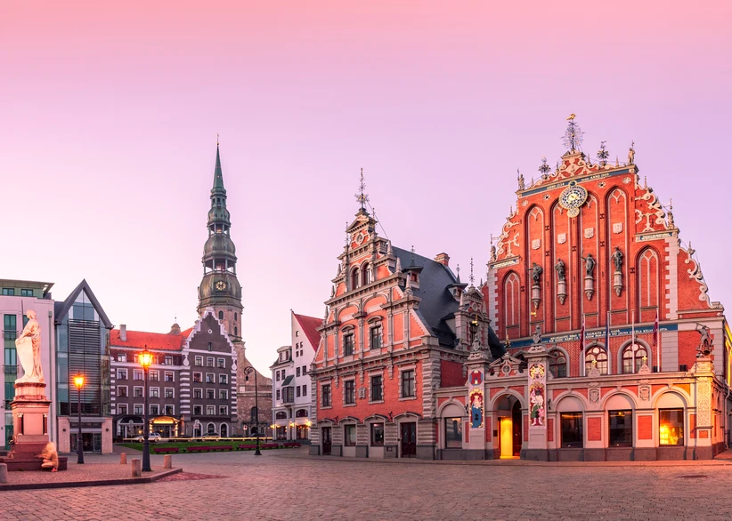 Riga's City Hall Square at dusk, with the ornate red-brick House of the Blackheads glowing under warm lights, St. Peter's Church spire in the background, and a statue and lamppost in the foreground under a pink sky.