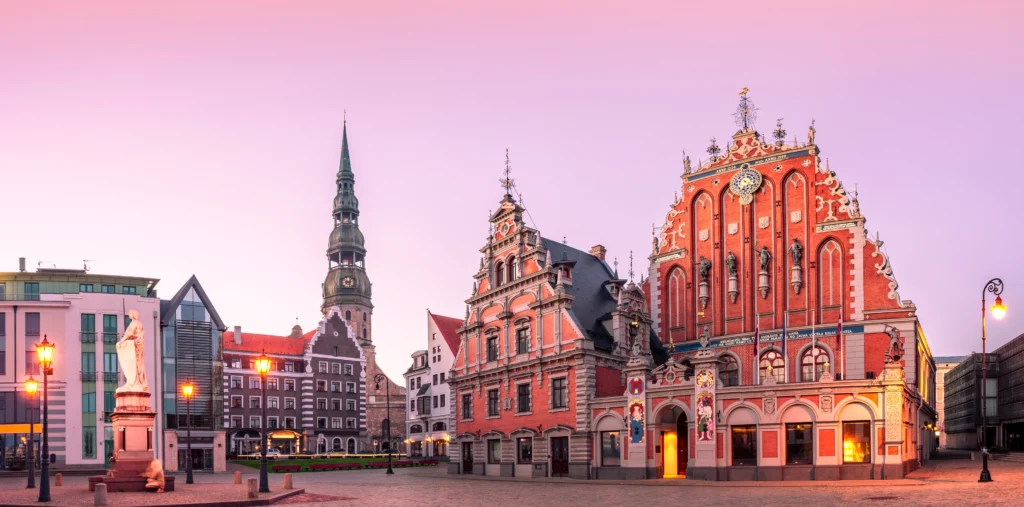 Riga's City Hall Square at dusk, with the ornate red-brick House of the Blackheads glowing under warm lights, St. Peter's Church spire in the background, and a statue and lamppost in the foreground under a pink sky.