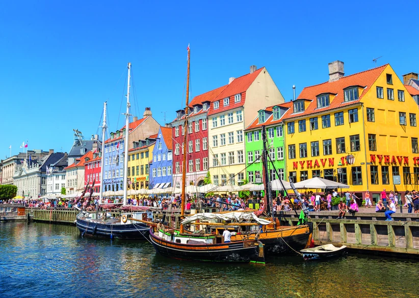 Colorful gabled townhouses in red, yellow, green, and blue line the sunny Nyhavn canal in Copenhagen, with wooden sailing ships moored along the waterfront and crowds filling the outdoor cafes on the quay.