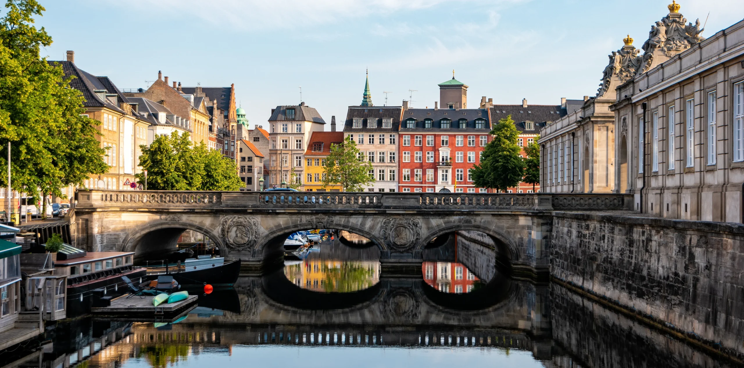 The ornate stone Marble Bridge crosses a canal toward Christiansborg Palace's crowned facade, with moored boats and colorful townhouses reflected in the still water and a green copper spire visible among the rooftops beyond.