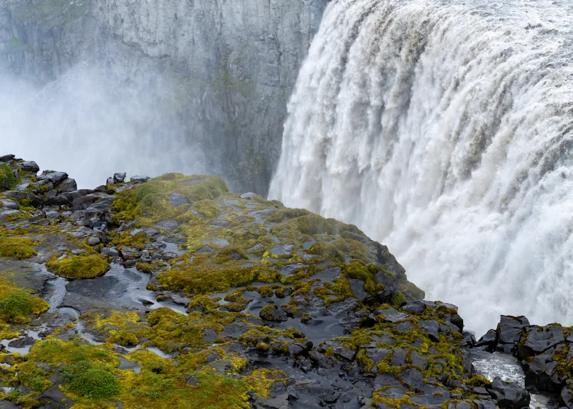 Powerful whitewater cascades over Dettifoss's basalt cliff into a misty canyon, with moss-covered black volcanic rocks lining the edge in the foreground.
