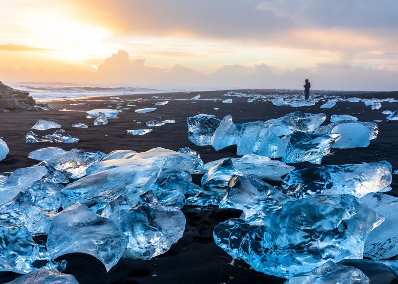 Large chunks of glacial ice glisten on the black volcanic sand of Diamond Beach, with a lone figure walking along the shoreline and a golden sunset glowing over the ocean in the background.