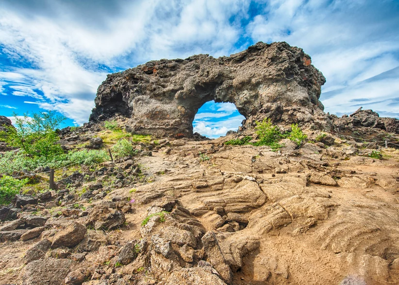 A dark, rugged lava rock arch with a natural opening rises above a rocky, moss-dotted trail at Dimmuborgir, under a bright blue sky with wispy clouds.