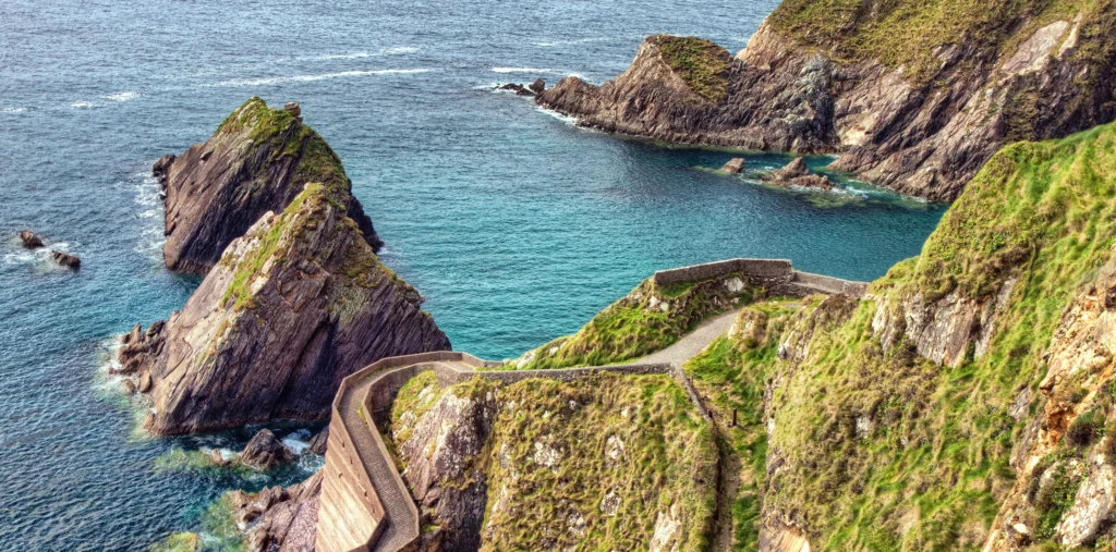 The winding stone path of Dunquin Pier descending through green cliffs past rugged sea stacks and turquoise Atlantic waters on the Dingle Peninsula, County Kerry, Ireland