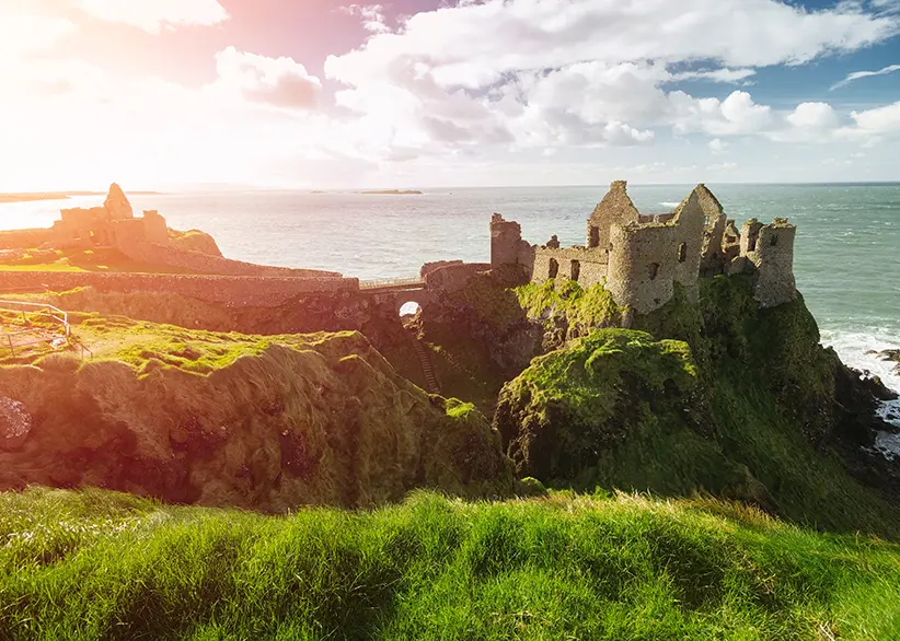 Dunluce Castle ruins perched on a dramatic cliff edge above the Atlantic Ocean with green grass, sunlight flaring through clouds, and waves crashing below, County Antrim, Northern Ireland