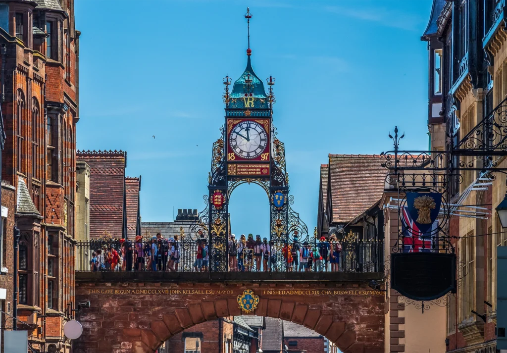 An ornate Victorian clock on a wrought-iron arch spans a sandstone gateway crowded with people below.