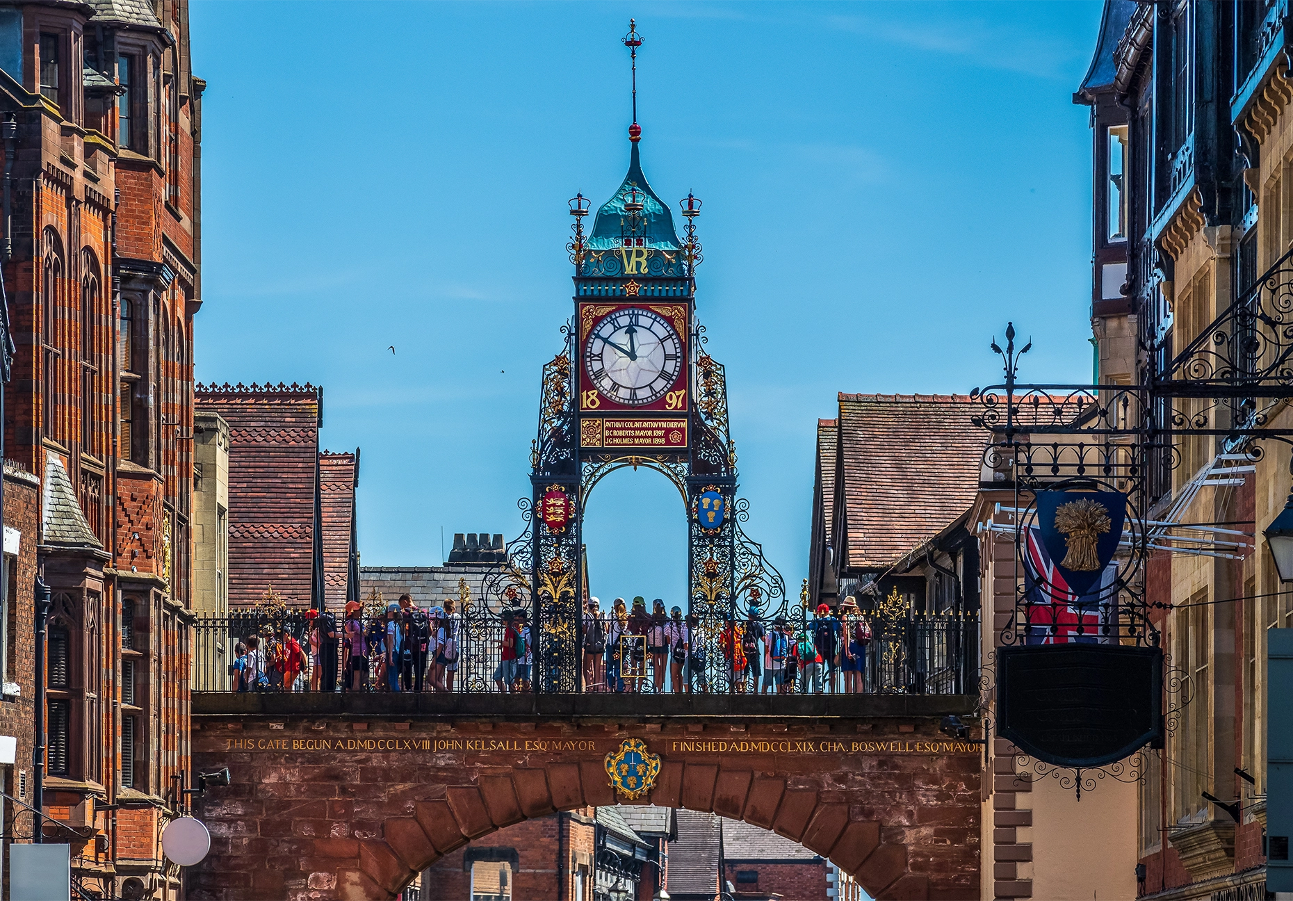 An ornate Victorian clock on a wrought-iron arch spans a sandstone gateway crowded with people below.