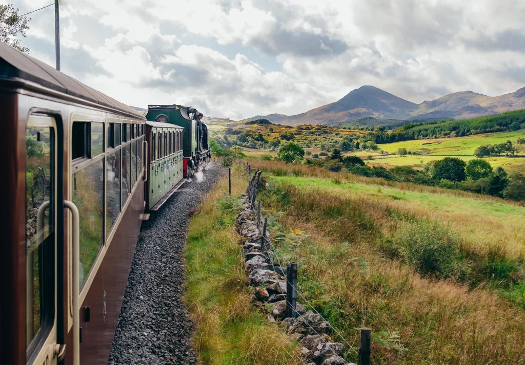 Heritage steam train with maroon carriages curves through a green valley below rugged peaks.