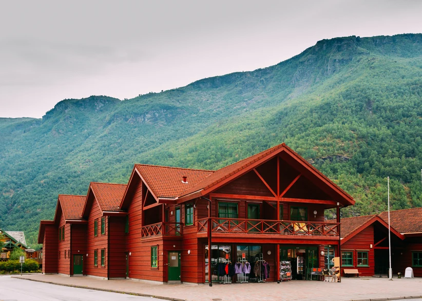 A large traditional red timber building with a balcony and gabled roofline stands before green, mist-covered mountains in Flåm, with outdoor clothing racks and a bench displayed outside its shop entrance.