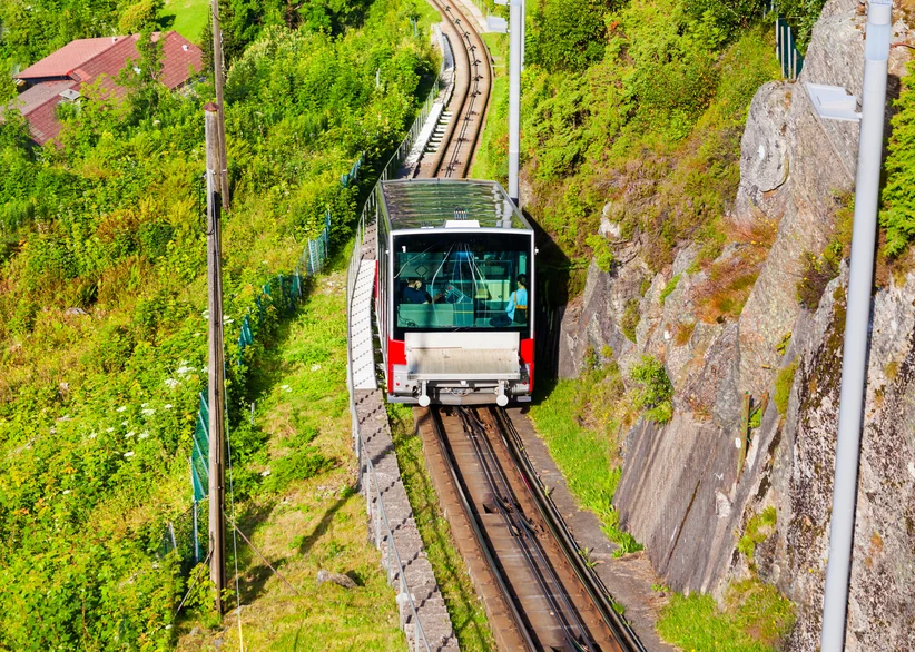 A red and white funicular car climbs a steep, forested hillside track cut through rock, with a red-roofed house and greenery visible alongside the rails in Bergen