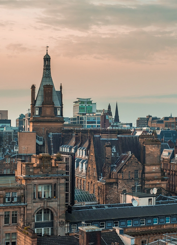 Historic Glasgow rooftops and a Victorian tower against a pink sunset sky.