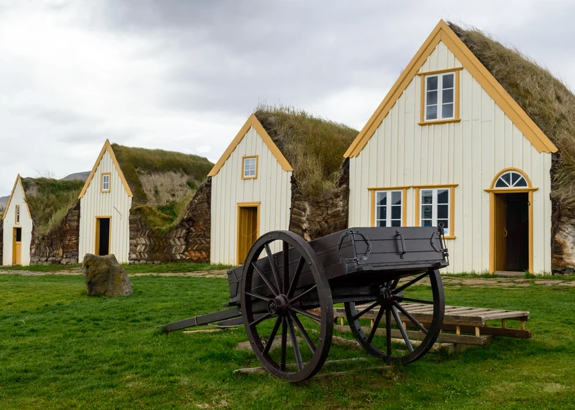 A row of traditional Icelandic turf houses with white gabled facades and grass-covered roofs at Glaumbær, with an old wooden cart parked on the grass in front.