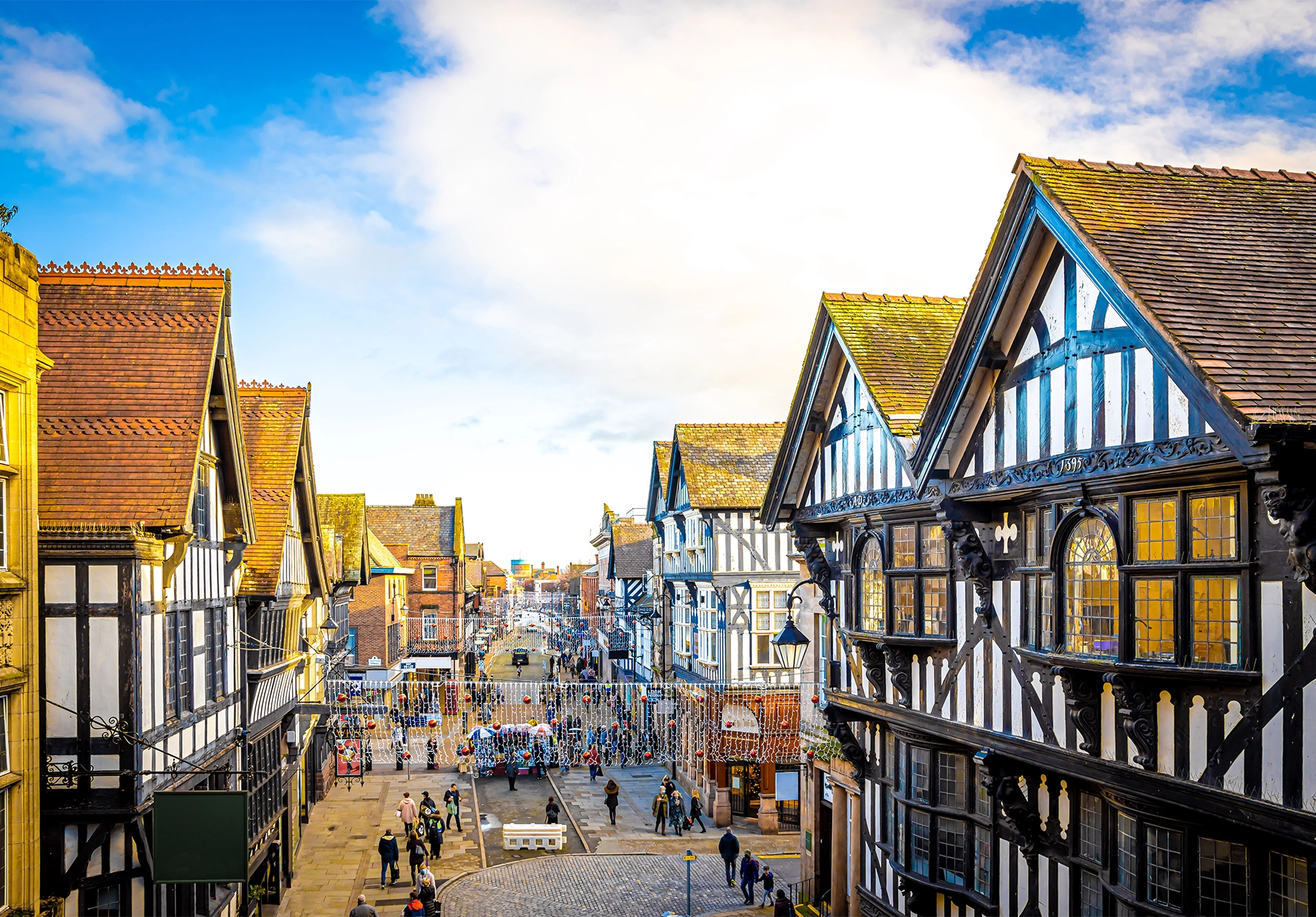 Elevated view of black-and-white timbered buildings lining a busy pedestrian shopping street under a blue sky.