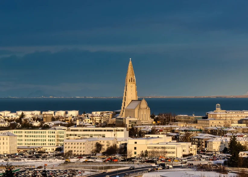 Hallgrímskirkja's tall, distinctive concrete spire rises above the snow-dusted rooftops of Reykjavik, with the ocean and distant coastline visible under a deep blue sky.