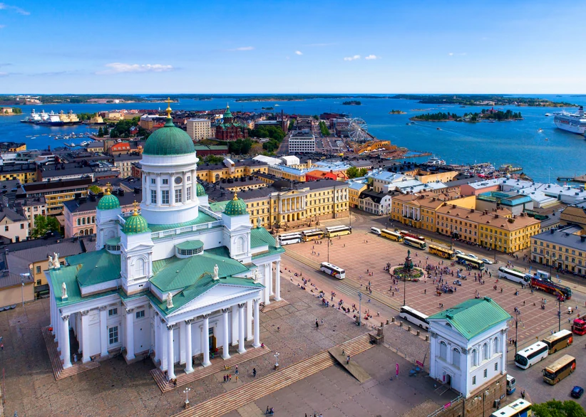 Aerial view of Helsinki Cathedral's white and green domes overlooking Senate Square, packed with tour buses and pedestrians, with the harbor, Ferris wheel, and scattered islands visible in the background.